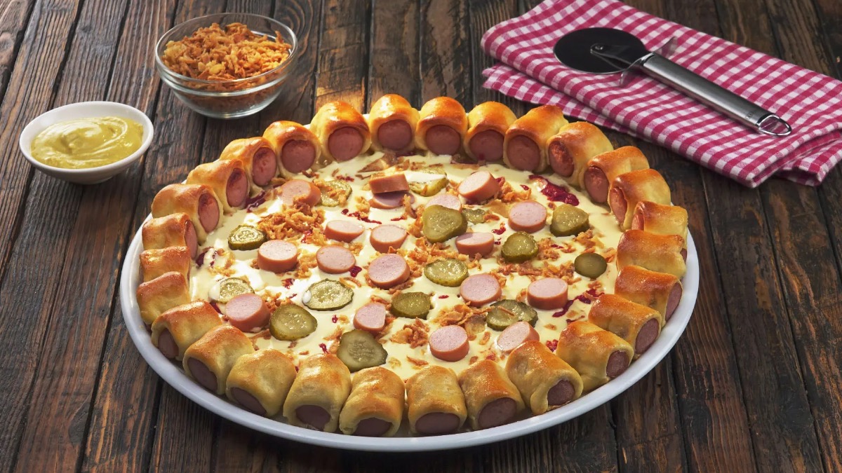Eine Hotdog-Pizza auf einem Teller, daneben eine Schale mit Röstzwiebeln, eine mit Senf und ein Pizzaschneider auf einem Tuch.