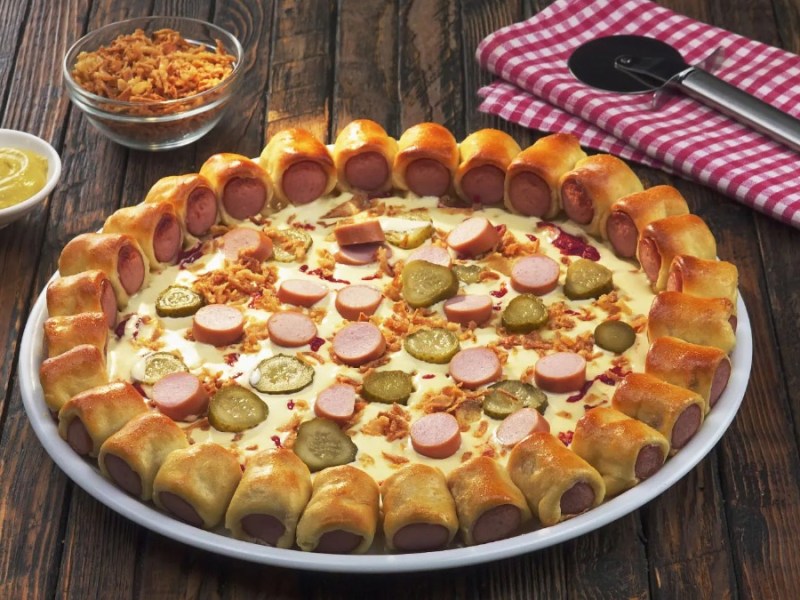 Eine Hotdog-Pizza auf einem Teller, daneben eine Schale mit Röstzwiebeln, eine mit Senf und ein Pizzaschneider auf einem Tuch.