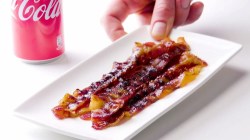 Eine Portion karamellisierter Bacon auf einem Teller wird auf den Tisch gelegt. Im Hintergrund steht eine Dose Cola.