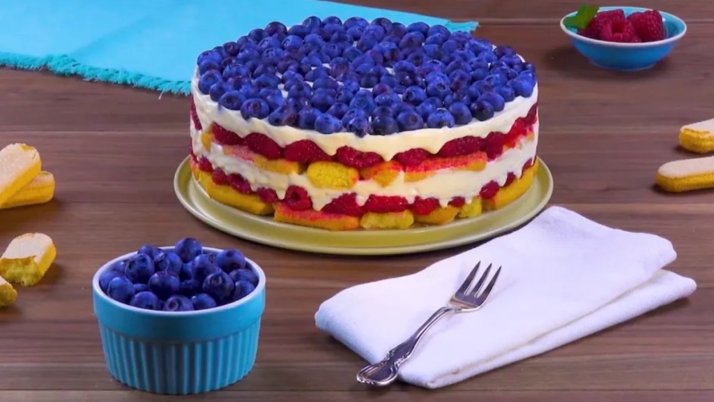 Eine bunte Löffelbiskuit-Torte mit einer Serviette mit einer Gabel und eine Schale mit frischen Blaubeeren daneben.
