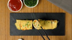 Eine japanische Omelett-Rolle auf einer Schieferplatte, aus der mithilfe von Essstäbchen ein Stück entnommen wird. Daneben zwei Schalen mit Dip und gehacktem Schnittlauch.