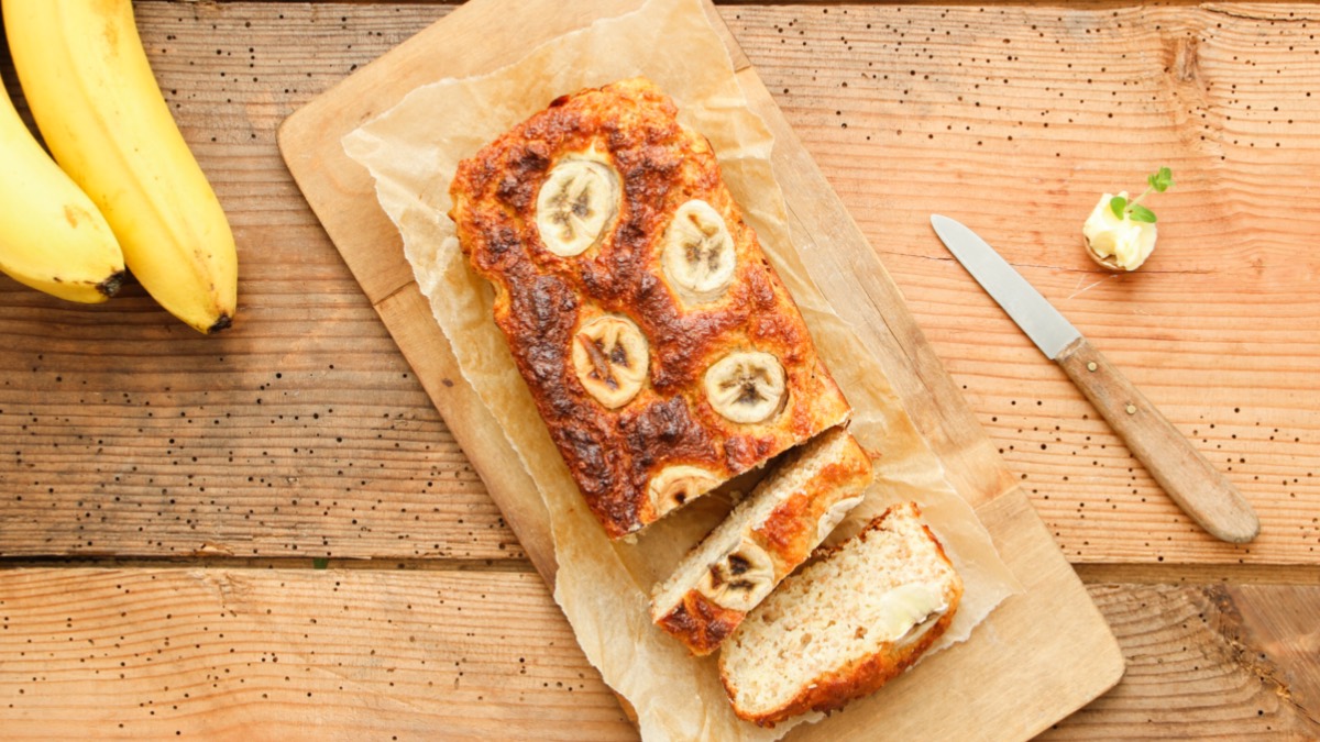 Angeschnittener Bananenkuchen auf Backpapier von oben fotografiert. Daneben liegen ein Messer und Bananen.