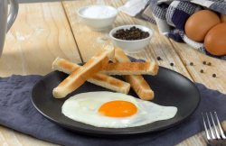 Ein Spiegelei aus der Mikrowelle auf einem schwarzen Teller mit Toast-Sticks. Im Hintergrund sind Eier und Schälchen im Pfeffer und Salz zu sehen.