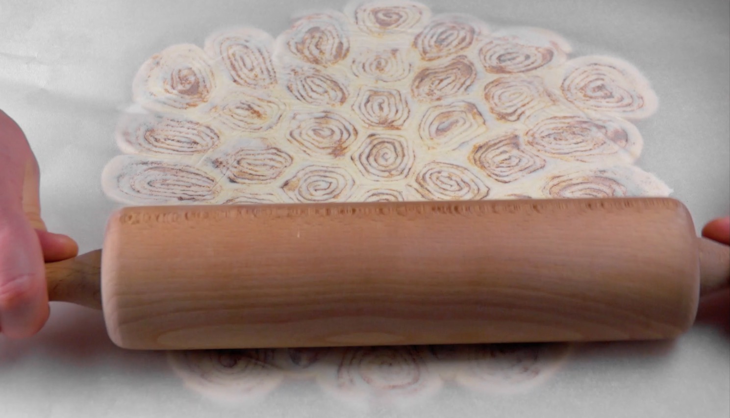 Mehrere aneinander gelegte Zimtschnecken werden mit einem Nudelholz plattgerollt