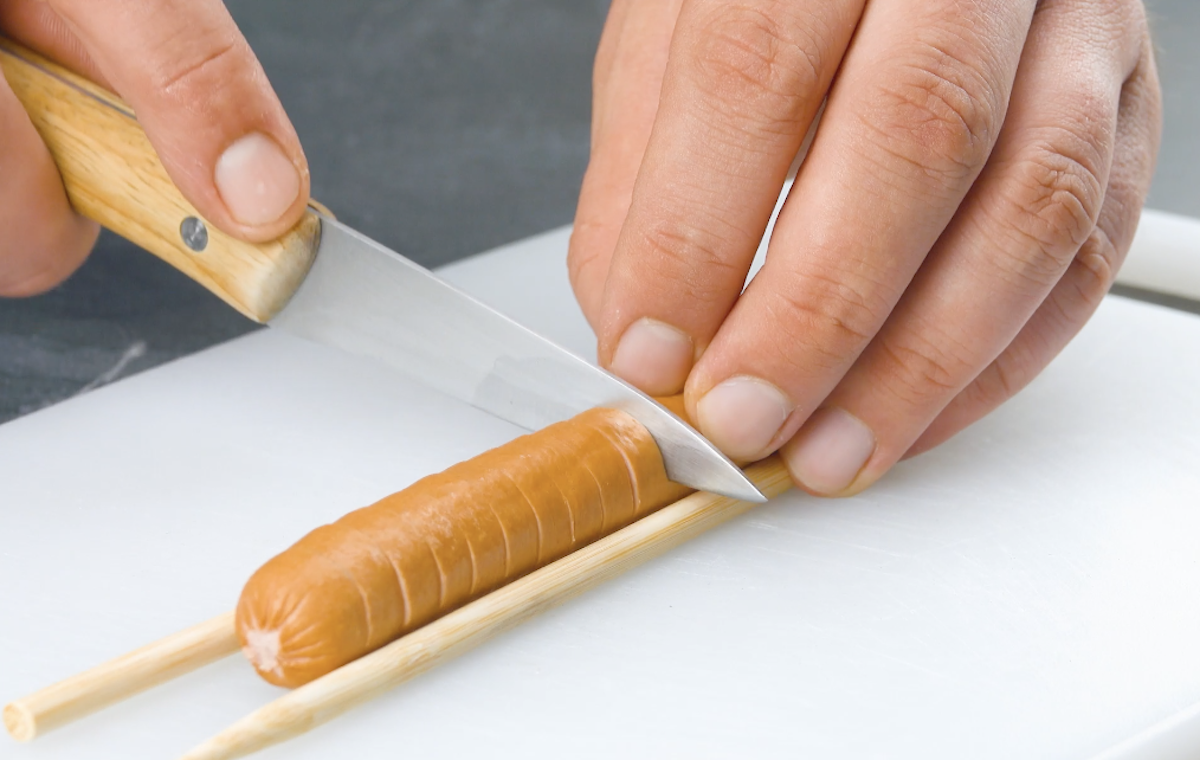 Ein Würstchen liegt zwischen zwei Holzstäben und wird mit einem Messer gleichmäßig eingeschnitten