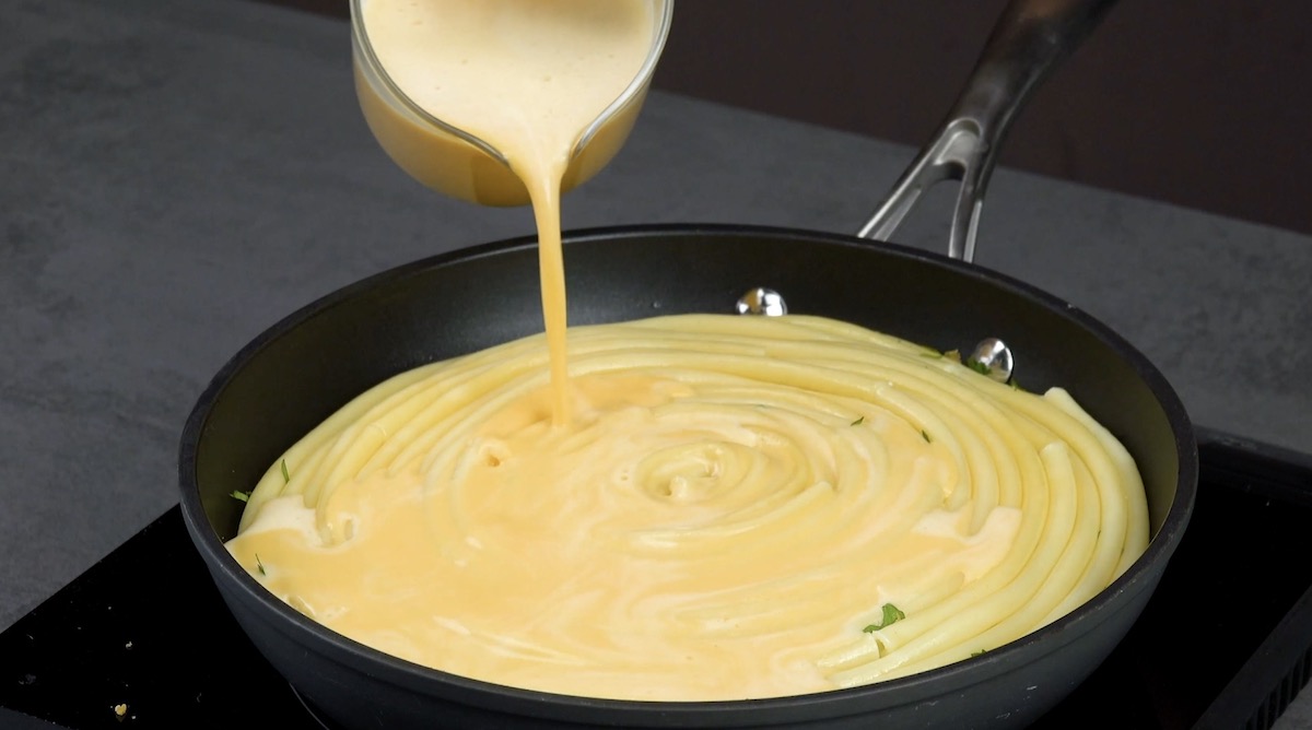 Mit Milch und Olivenöl verquirlte Eier werden über Nudeln und Käse in Pfanne gegossen