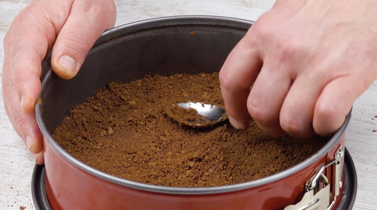 Zerbröselte Butterkekse werden mit Löffel als Boden in Kuchenform gedrückt