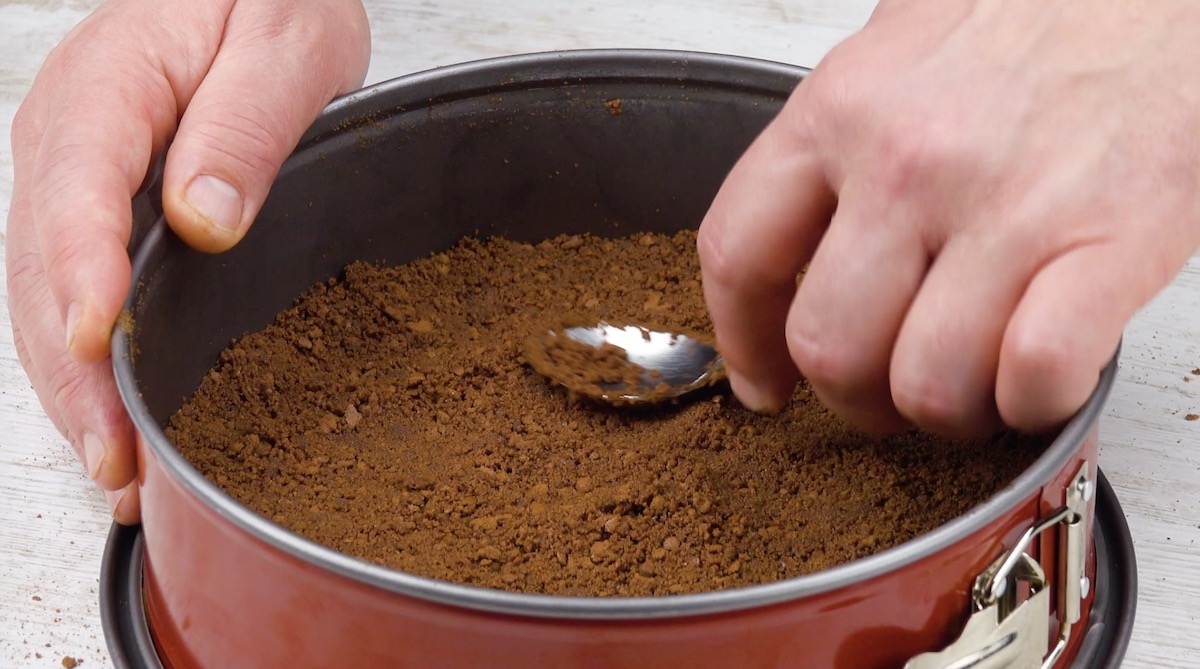 ZerbrÃ¶selte Butterkekse werden mit LÃ¶ffel als Boden in Kuchenform gedrÃ¼ckt