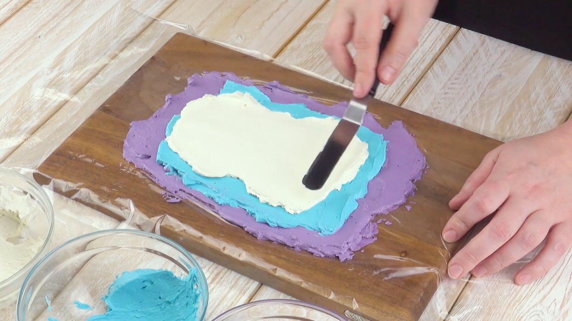 Buttercremes in Lila, Blau und WeiÃŸ werden Ã¼bereinander auf Frischhaltefolie gestrichen