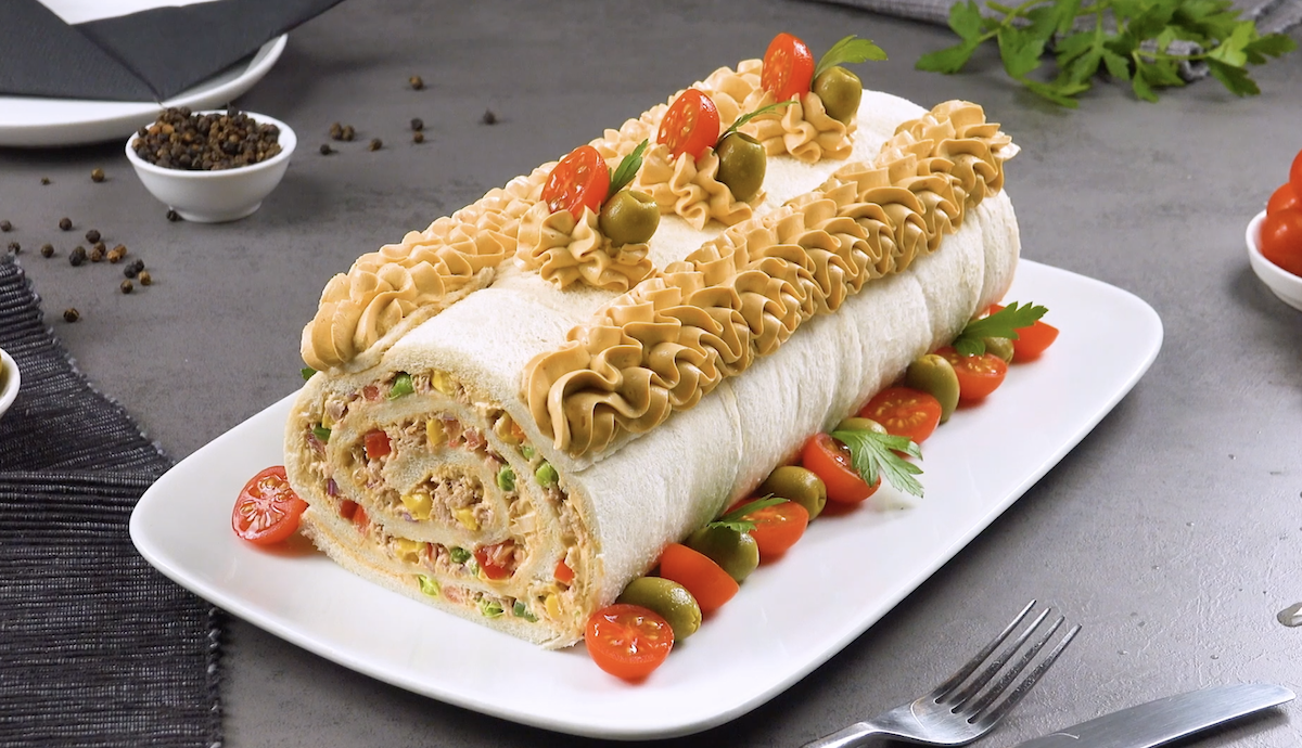 Eine Thunfisch-Sandwich-Rolle auf einer länglichen Servierplatte mit Tomaten und Oliven als Dekoration.