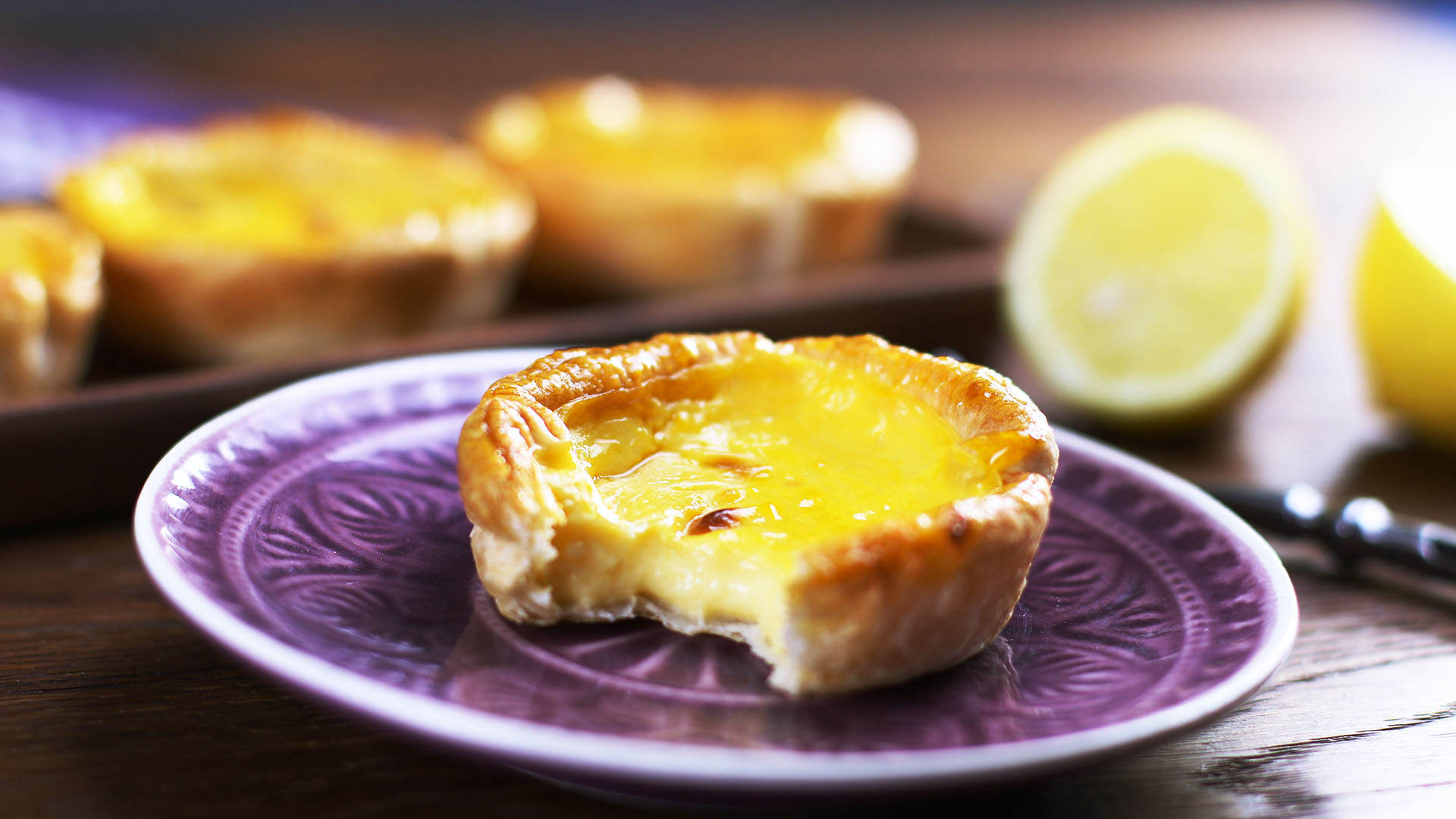 Ein angebissenes portugiesisches Puddingtörtchen mit weiteren Törtchen und einer halbierten Zitrone im Hintergrund.