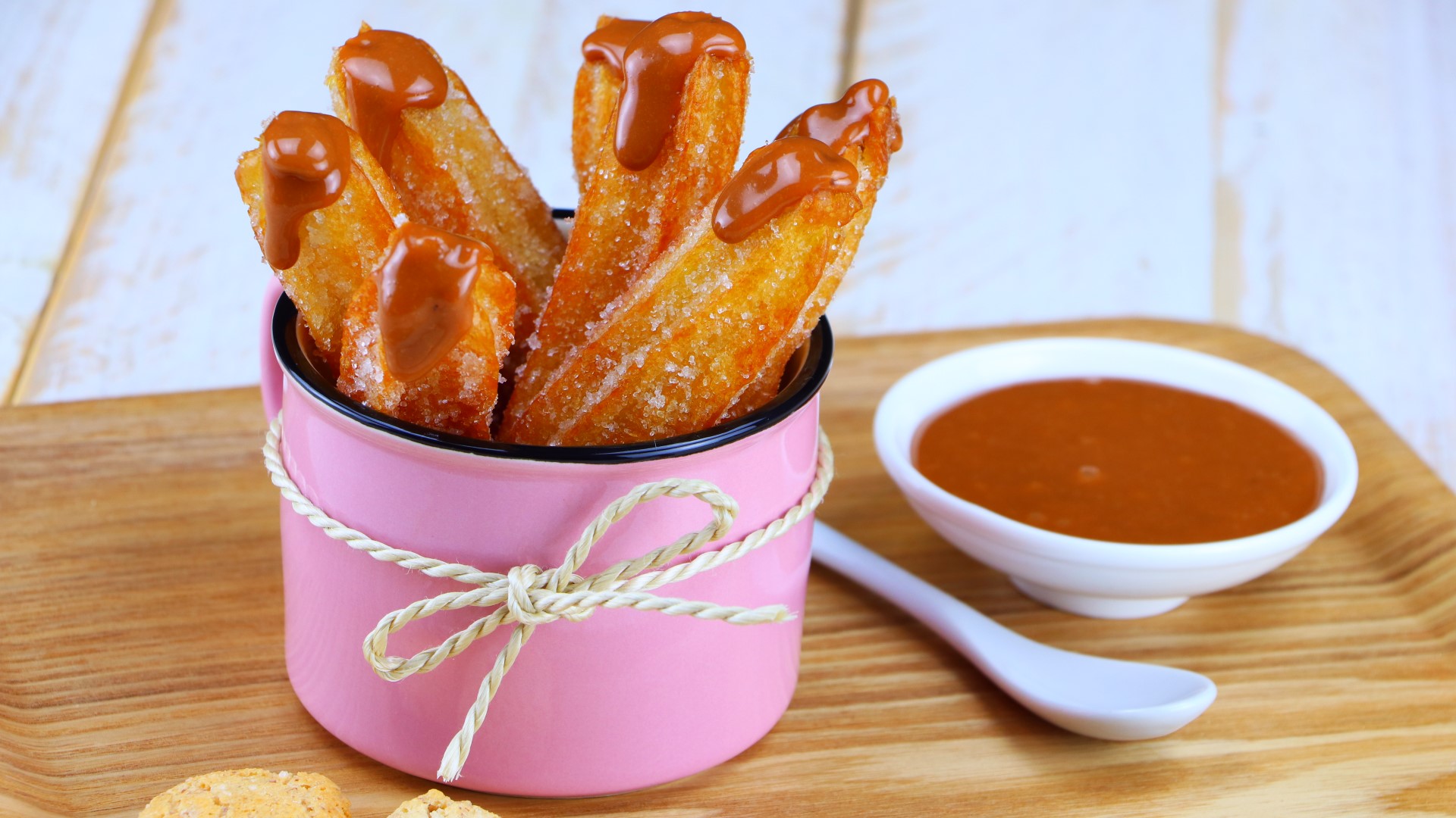 Churros mit Karamell-Creme in einer Tasse, daneben ein Löffel und eine Schale mit Karamell-Creme.