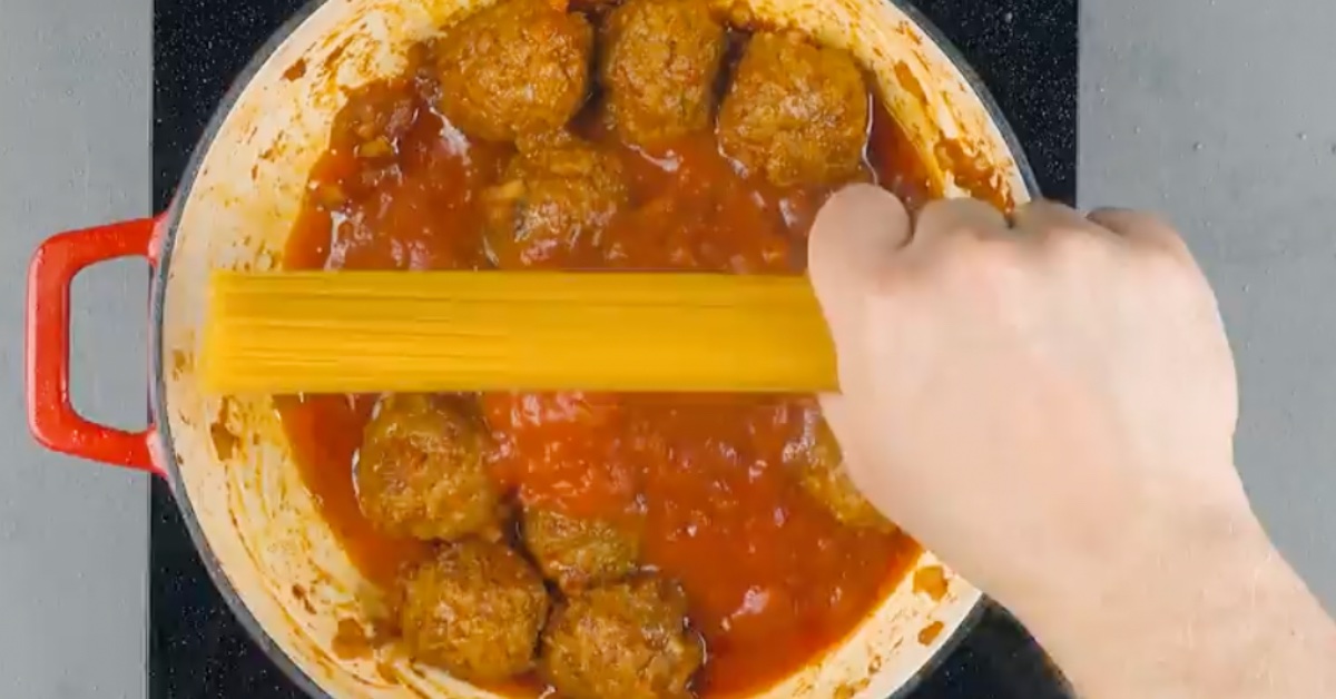 Eine Hand hält spaghetti über einen Topf mit Tomatensoße und Hackfleischbällchen.
