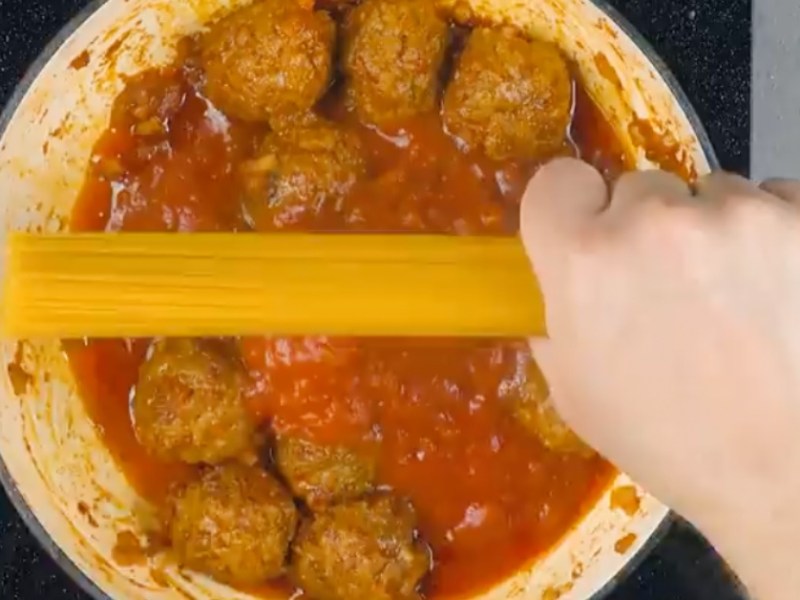 Eine Hand hält spaghetti über einen Topf mit Tomatensoße und Hackfleischbällchen.