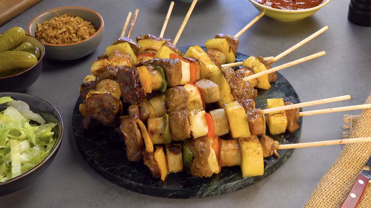 Selbstgemachte Grillspieße aus Hackfleisch, Gemüse und Ananas auf einem Teller, daneben Schälchen mit Zutaten.