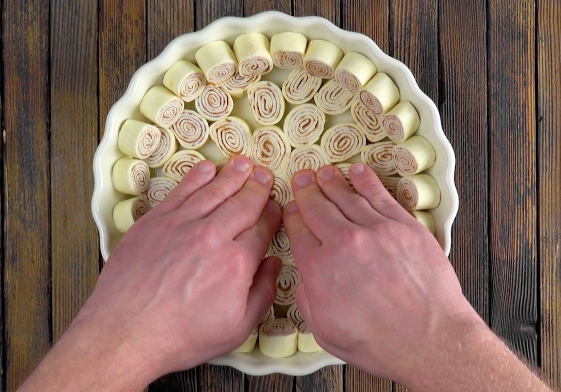 Mehrere kleine Zimtschnecken werden mit zwei Händen auf den Boden einer Kuchenform gedrückt