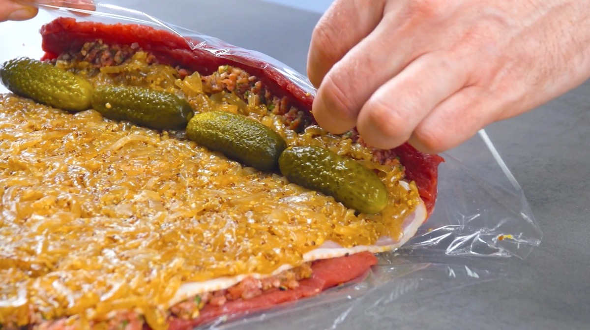 Große Rinderroulade mit Essiggurken auf karamellisierten Zwiebeln und Bacon wird in Frischhaltefolie eingerollt