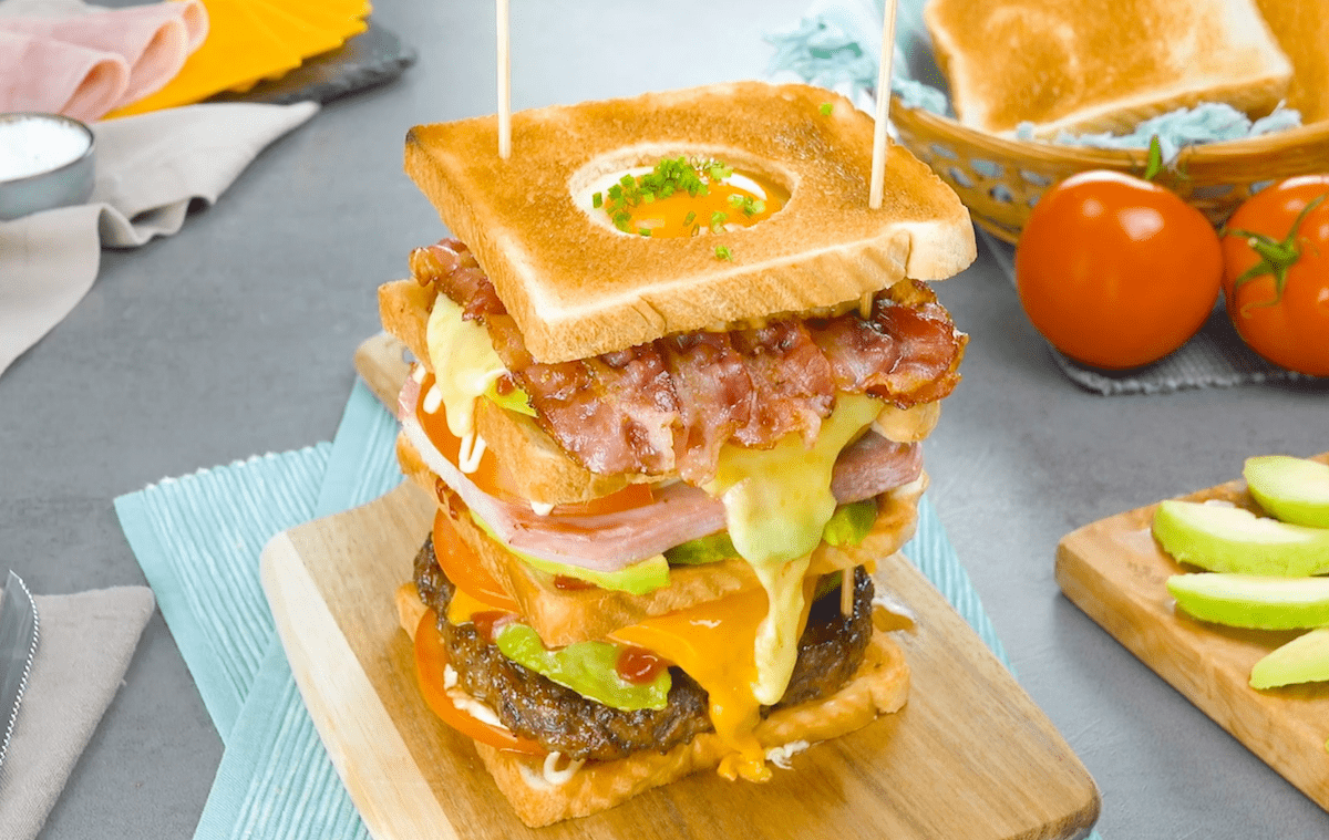 Ein Sandwich-Turm auf einem Holzbrett. Im Hintergrund frische Tomaten und ein Brotkorb mit Toasts. 
