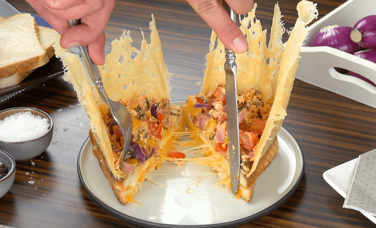 Ein Toastbrot mit Käsekrone auf einem Teller wird halbeirt und mithilfe von Besteck auseinander gezogen. 
