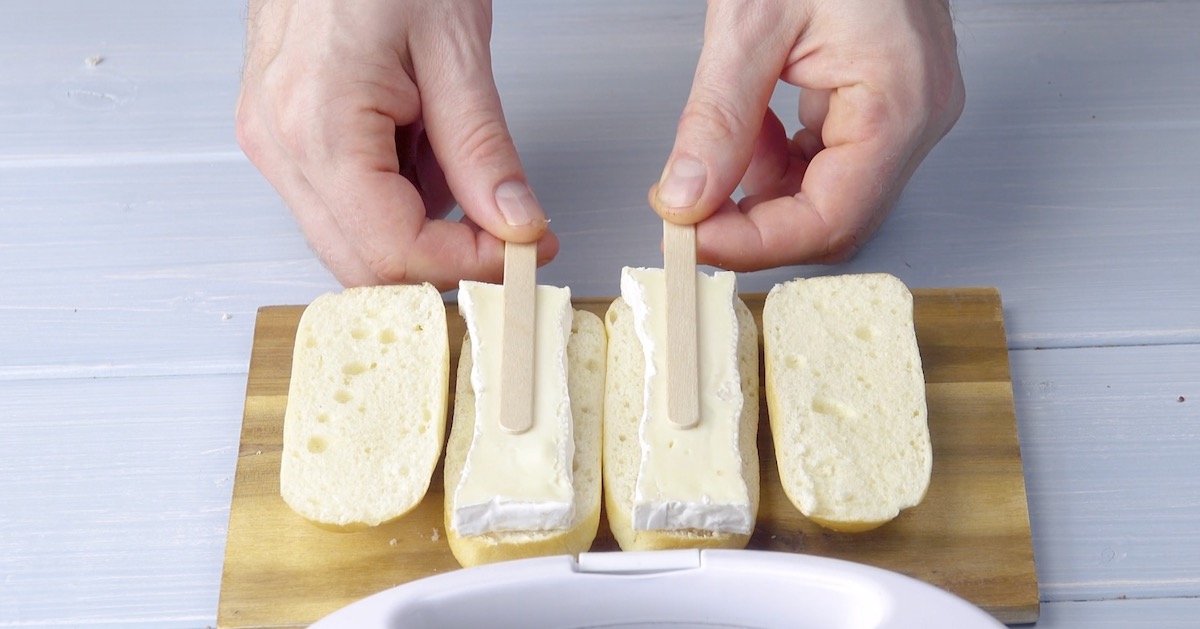 Auf zwei MilchbrÃ¶tchenhÃ¤lften werden je eine Scheibe Camembert und ein Eisstiel platziert.