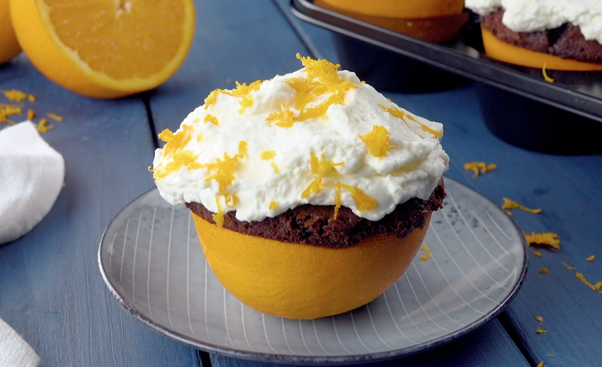 Ein Muffin in der Orangenschale auf einem Teller. Im Hintergrund ist eine aufgeschnittene Orange und das Muffinblech mit weiteren Muffins zu sehen.