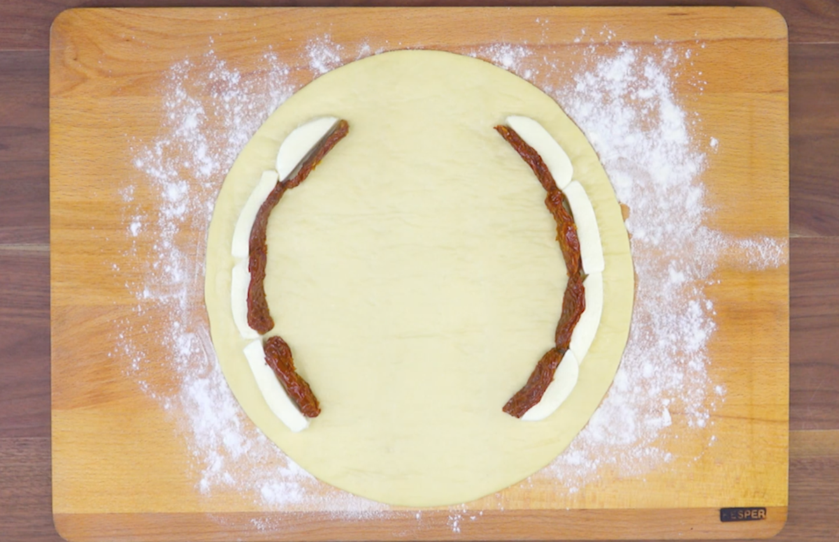 Pizzateig wird auf Brett ausgerollt und Mozzarella und getrocknete Tomatenscheiben klammerfÃ¶rmig darauf gelegt