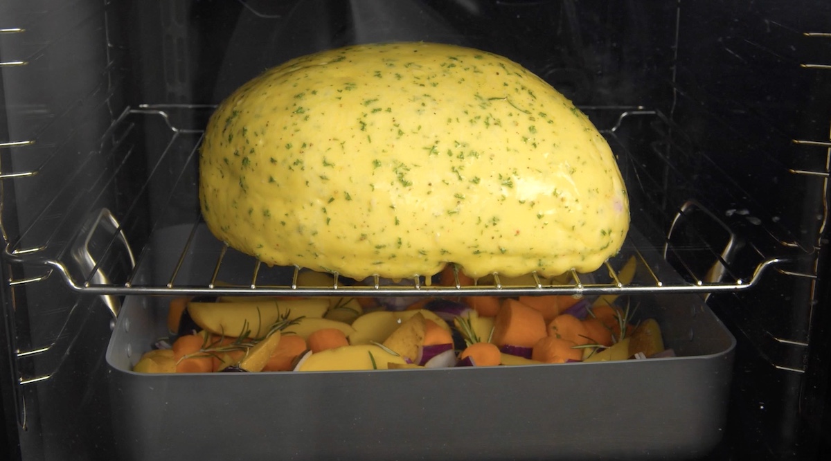 Mit Kräuterbutter bestrichenes Hühnchen liegt auf Gitterrost im Oden über einer Auflaufform mit Gemüse