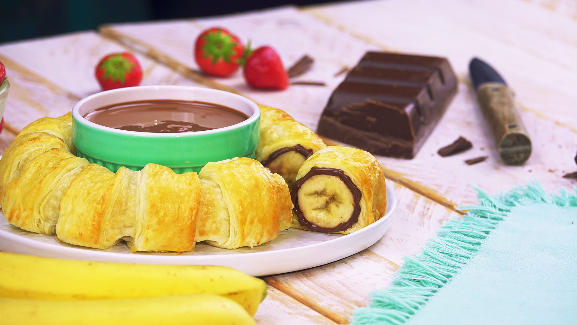 Ein Nutella-Bananen-Ring mit einer Schale Nutella in der Mitte, daneben Erdbeeren, Bananen und ein Block Kuvertüre.