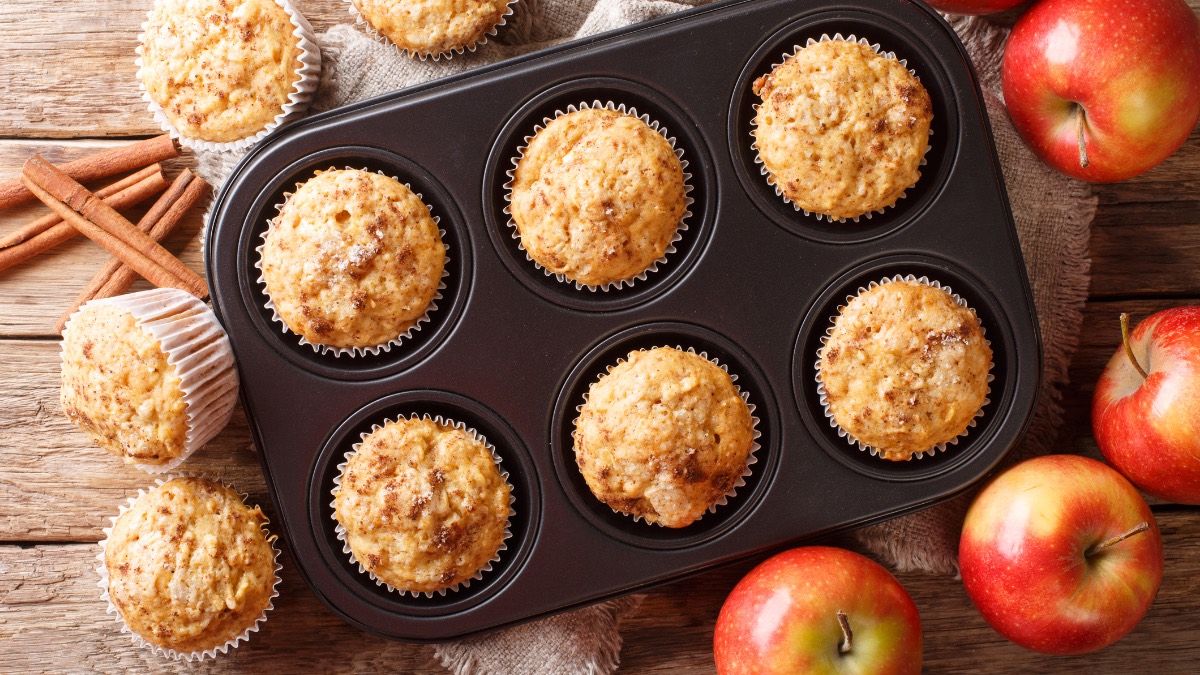 Ein Muffinblech mit Apfel-Zimt-Muffins. Daneben liegen Äpfel und weitere Muffins.