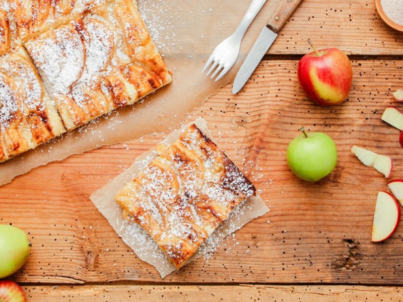 Ein Stück Apfelkuchen vom Blech mit Puderzucker. Daneben Äpfel, Besteck und der restliche Kuchen.