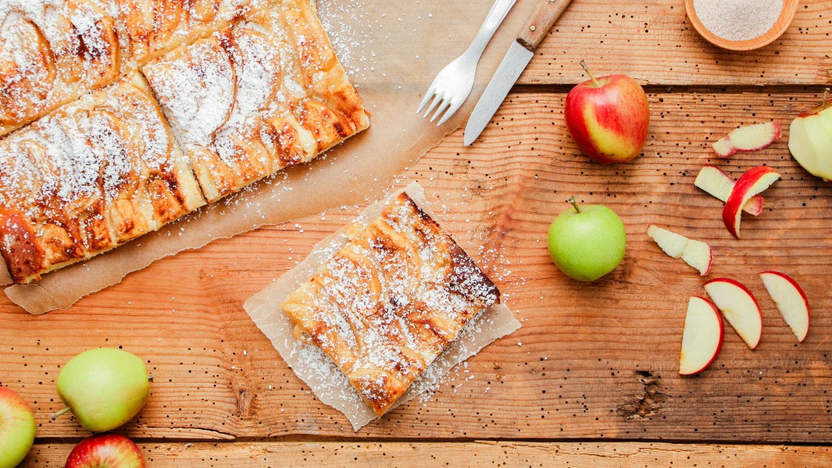 Ein Stück Apfelkuchen vom Blech mit Puderzucker. Daneben Äpfel, Besteck und der restliche Kuchen.