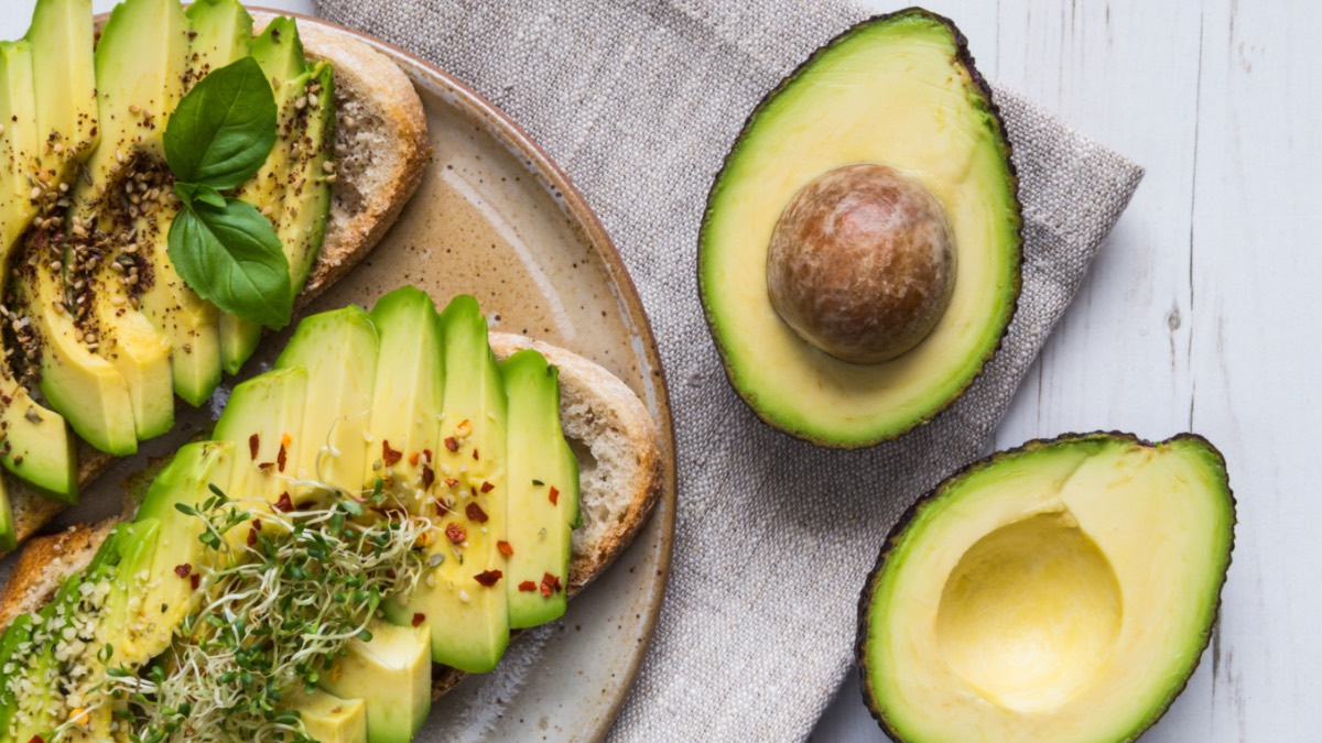 Eine halbierte Avocado neben zwei Broten mit Avocado auf einem Teller.