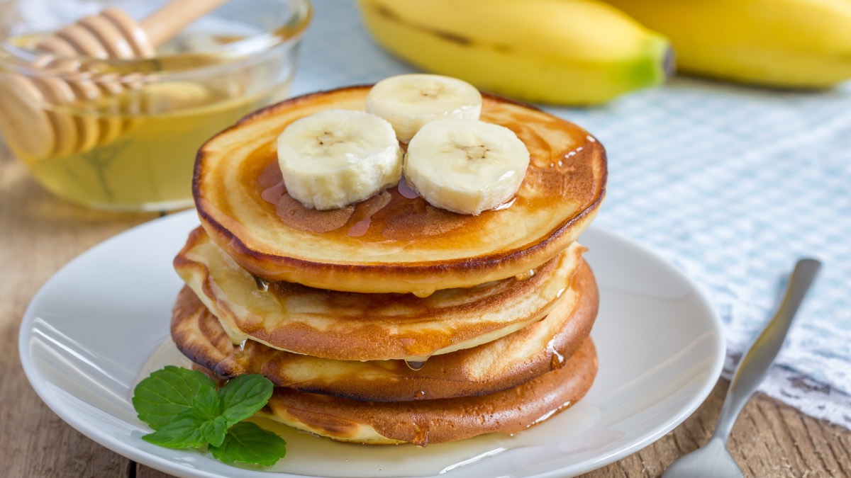 Ein Stapel Bananen-Pancakes mit Ahornsirup und Bananenscheiben. Im Hintergrund liegen Bananen.