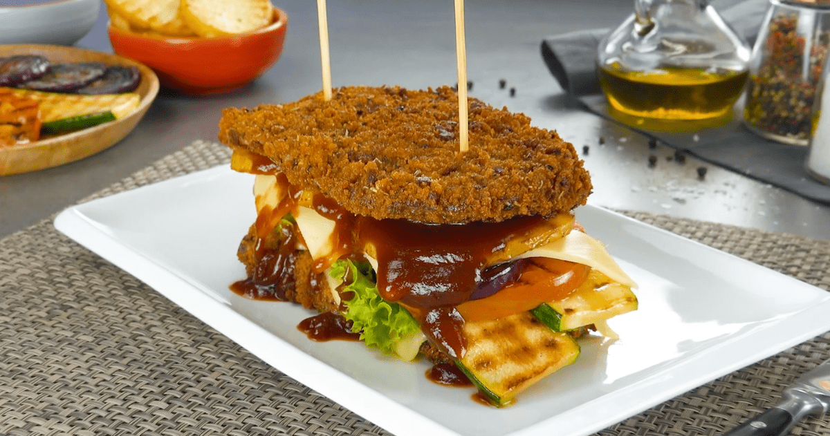Ein Rumpsteak-Sandwich mit Panade aus Graubrot auf einem Teller. Daneben eine Schale und ein Teller mit den Zutaten und eine Karaffe mit Öl und Salz- und Pfeffermühle.