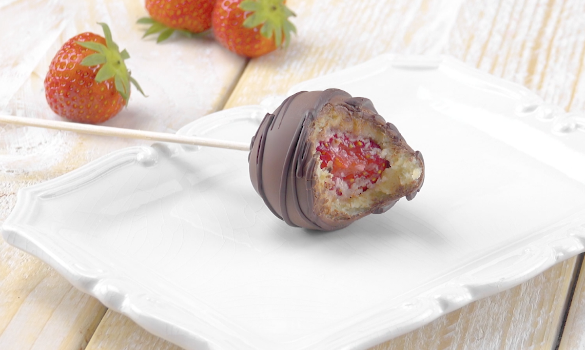 Eine mit Schokolade überzogene Erdbeere am Spieß, die angebissen auf einem Teller liegt. Im Hintergrund liegen drei frische Erdbeeren. 