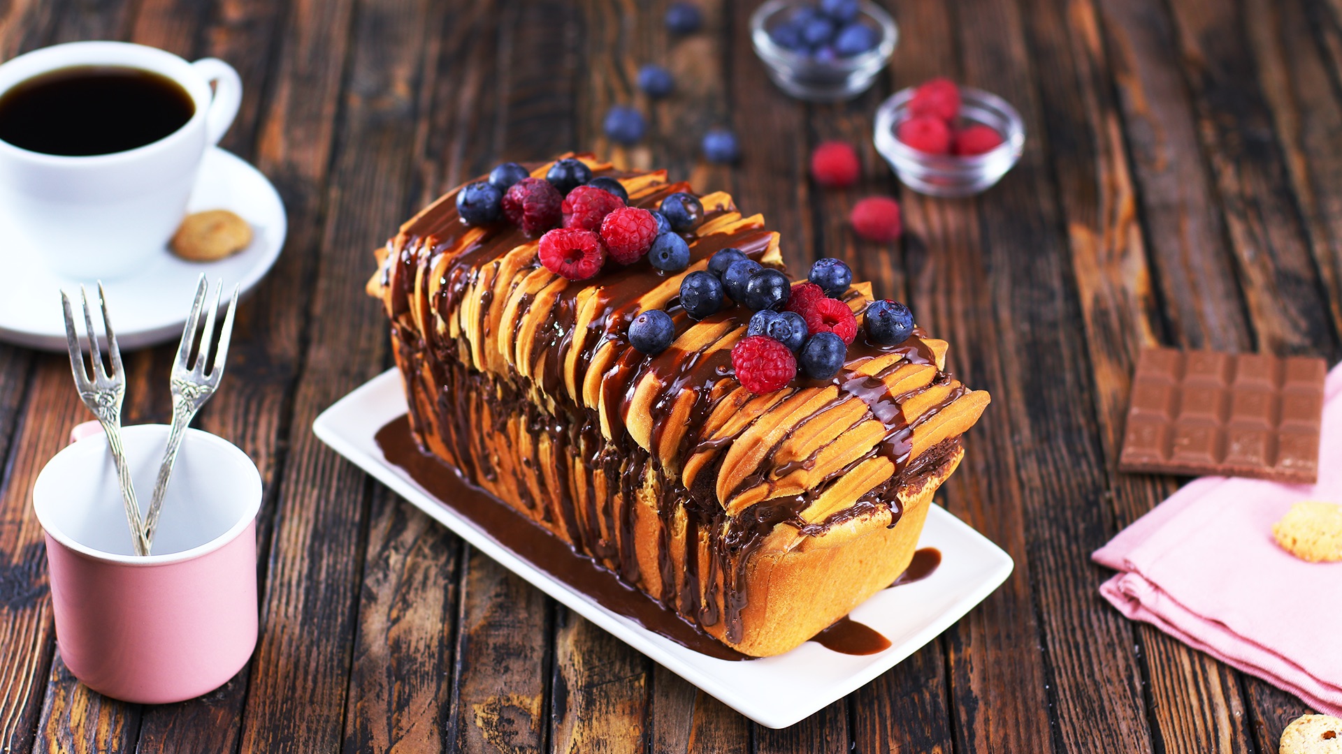 Ein Ziehharmonika-Kuchen mit Nutella, daneben eine Tasse Kaffee, eine Tasse mit Gabeln und frische Beeren und eine halbe Tafel Schokolade.