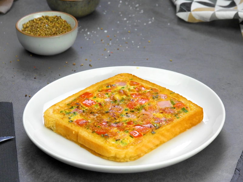 Ein Pizza-Toast auf einem Teller, drumherum Zutaten und Küchenutensilien.