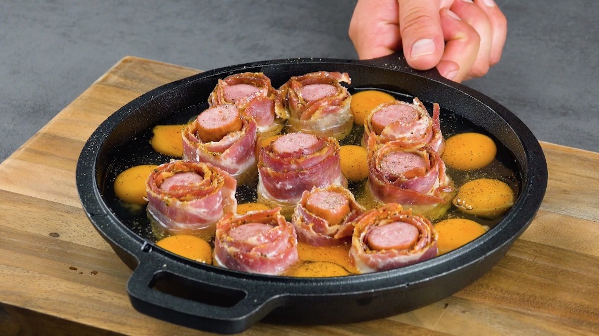 WÃ¼rstchen mit Bacon werden in Pfanne platziert und rohe Eier hinzugefÃ¼gt