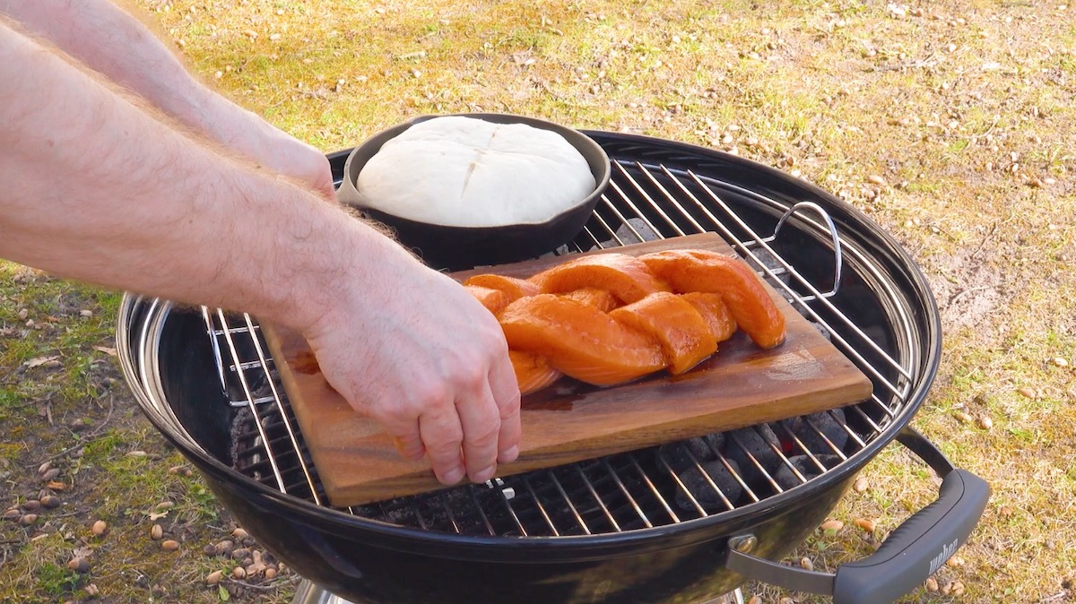 Lachszopf auf Grillbrett kommt neben Brot in Pfanne auf dem Grill