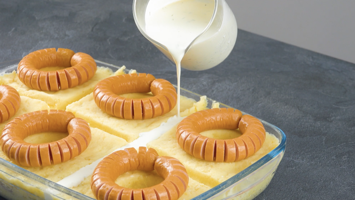 Sahne wird in eine Auflaufform mit Kartoffelstampf und Würstchen gegeben