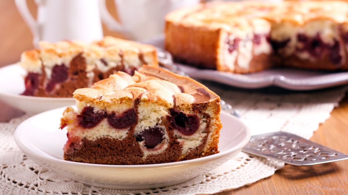 Ein Stück gefüllter Marmorkuchen mit Kirschen auf einem Teller. Im Hintergrund sind der restliche Kuchen und ein weiteres Stück auf einem Teller zu sehen.