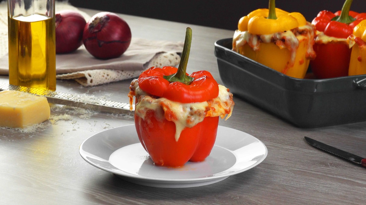 Eine Mini-Lasagne in Paprika auf einem Teller. Im Hintergrund sind weitere gefüllte Paprikaschoten und Zutaten zu sehen. 