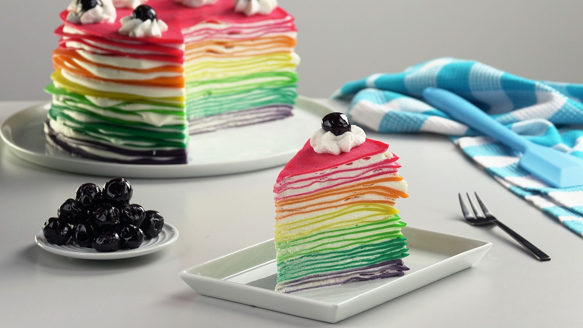 Teller mit einem Stück Regenbogen-Crêpe-Kuchen und einer angeschnittenen Torte im Hintergrund