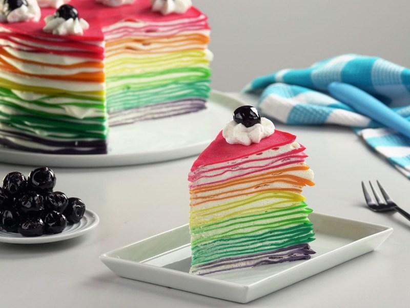Teller mit einem Stück Regenbogen-Crêpe-Kuchen und einer angeschnittenen Torte im Hintergrund