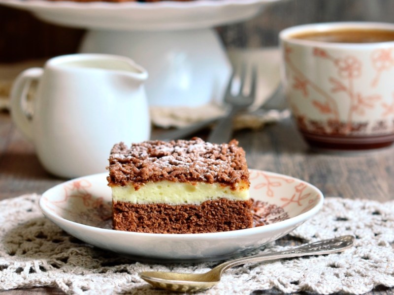 Ein Stück Schoko-Streuselkuchen, daneben eine Tasse Kaffee, ein Kännchen Milch und der restliche Kuchen.