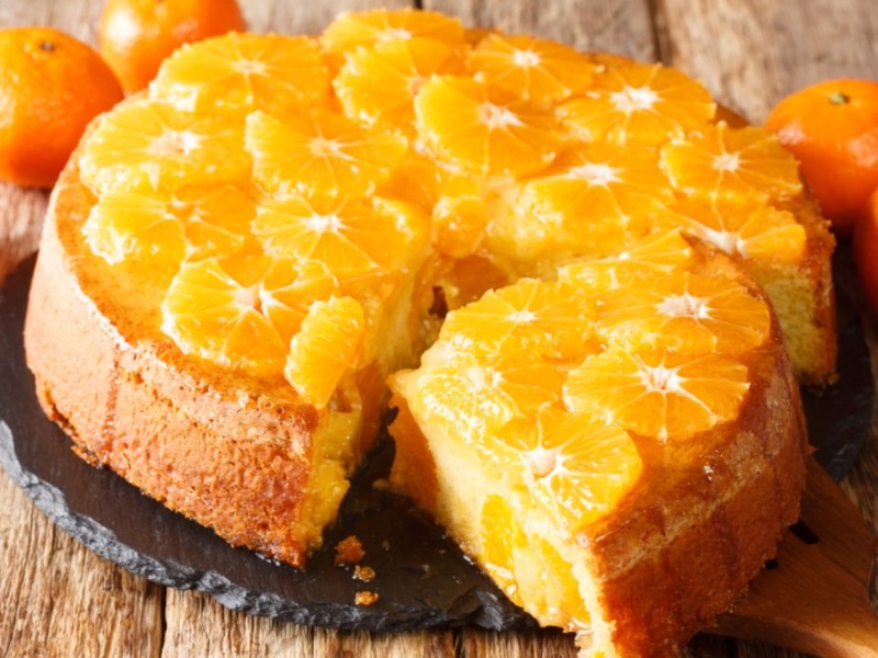 Ein Schüttelkuchen mit Mandarine. Ein großes Stück wurde herausgeschnitten und liegt vor dem Kuchen.