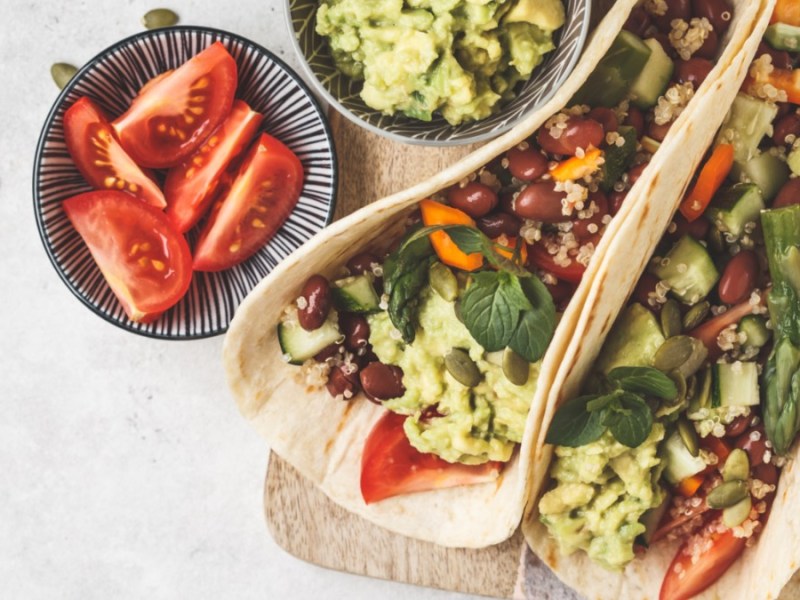 Zwei Spargel-Wraps mit Quinoa und Guacamole nebeneinander. Daneben stehen Schälchen mit Zutaten.