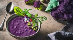 Lilafarbene Süßkartoffel-Blumenkohl-Suppe mit Salat-Dekoration. Daneben liegt ein Löffel und im Hintergrund Zutaten.