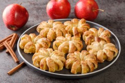 Mehrere Apfel-Blätterteig-Donuts auf einem Teller, daneben frische Äpfel und Zimtstangen.