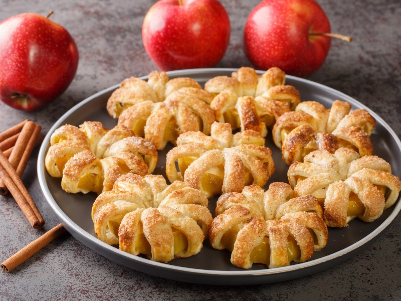 Mehrere Apfel-Blätterteig-Donuts auf einem Teller, daneben frische Äpfel und Zimtstangen.
